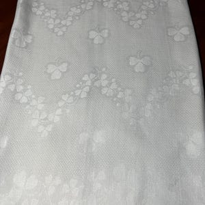 Può includere: Un tessuto bianco caratterizzato da un motivo damascato intrecciato con motivi ripetuti di trifogli. Il design include una prominente fascia a zigzag di piccoli trifogli al centro, con forme di trifoglio individuali più grandi sparse.