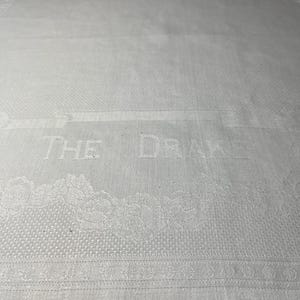 May include: A white rectangular textile featuring a woven damask pattern. The fabric displays the text THE DRAKE in a prominent font, surrounded by decorative floral borders and intricate scrollwork details integrated into the weave.