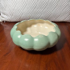 May include: A round, shallow ceramic bowl with a scalloped, pumpkin-like edge. The exterior features a matte, light mint green finish, while the interior is a smooth, glossy off-white color. The bowl has a wide, open top and a curved base.