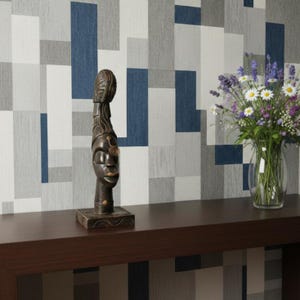 Könnte beinhalten: Eine dunkelbraune Holzskulptur eines Gesichts mit detailliertem Kopfschmuck steht auf einem dunkelbraunen Tisch. Eine klare Glasvase mit einem Strauß weißer Gänseblümchen und lila Blumen befindet sich auf der rechten Seite. Die Wand hat ein geometrisches Muster aus blauen, grauen und weißen Rechtecken.