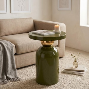 Può includere: Un tavolino verde oliva lucido con base cilindrica e un dettaglio sferico dorato che collega il piano rotondo. Un libro bianco e una candela a colonna bianca su un piattino poggiano sulla superficie del tavolo, caratterizzata da una finitura liscia.