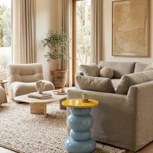 May include: A side table featuring a glossy yellow circular top and a stacked, bulbous light blue base. The table stands on a textured rug in a living room setting, positioned next to a gray sofa and a beige armchair with a coffee table.