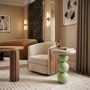 May include: A side table featuring a smooth, light beige circular top supported by a vertical stack of three glossy, pale green spherical spheres. The table is positioned next to a cream-colored upholstered armchair with dark wood side panels.