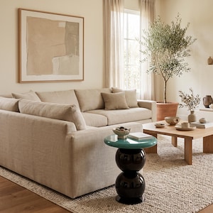 Puede incluir: Una mesa auxiliar con una superficie circular de color verde azulado brillante y una base esférica negra apilada, situada junto a un sofá tapizado en color beige. La mesa presenta un acabado liso y reflectante en la parte superior.