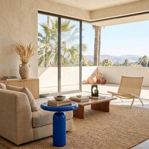 May include: A vibrant blue side table with a cylindrical base and a round top, positioned next to a beige upholstered sofa. The table features a smooth, glossy finish and stands on a textured brown rug in a room with large glass sliding doors.