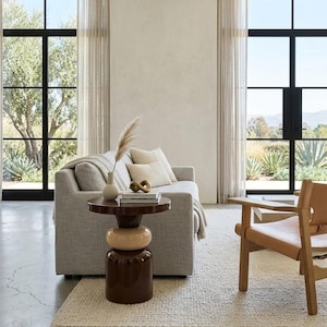 May include: A light grey sofa with a textured throw blanket and two beige cushions, positioned next to a round, dark brown and cream stacked-form side table. A tan leather armchair sits nearby on a cream rug in a room with large black-framed windows.