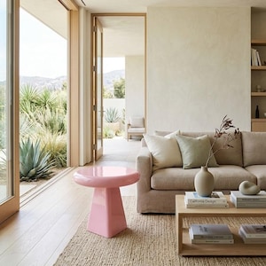 May include: A glossy, light pink side table with a circular top and a tapered, square-based pedestal. The table features a smooth, reflective finish and stands independently on a light-colored wooden floor, positioned near a beige sofa and rug.