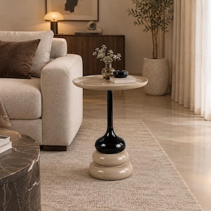 Puede incluir: Una mesa auxiliar con un tablero redondo de color beige, sostenido por una columna central con una sección negra brillante y una base beige apilada y redondeada. El diseño presenta líneas suaves y una paleta de dos colores.