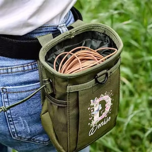 Peut inclure: Une pochette utilitaire vert olive avec un "D" floral et le nom "Denise" imprimé sur le devant. La pochette est attachée à une ceinture noire et contient une corde marron enroulée. La pochette a un anneau en D noir et un fermoir noir.