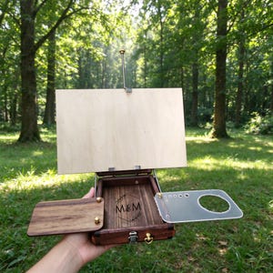May include: A portable wooden art easel box with a dark wood finish, featuring a light wood panel, a fold-out side tray, and a clear acrylic palette with circular cutouts. The interior base displays the engraved letters M&M inside a diamond shape.