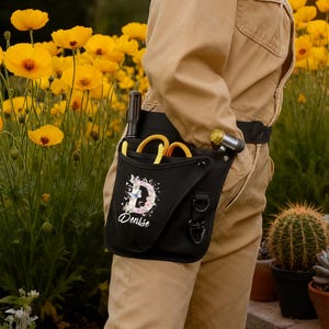 May include: Black gardening tool belt with a floral "D" design and the name "Denise". The belt holds gardening tools, including yellow handled shears and a small mallet. The background features yellow flowers.