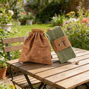 May include: Two waxed canvas pouches on a wooden table. One is brown with a drawstring closure. The other is olive green with a leather strap that reads "Janice". The background features a garden with flowers and greenery.