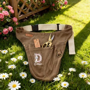 May include: A brown canvas gardening tool belt with a beige adjustable strap and black buckle. The belt has pockets, a gold-coloured pair of scissors, and a tag. The belt is personalised with the name "Denise" and a floral design.