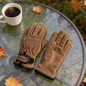 May include: Brown suede gloves with black trim and a black wrist strap, next to a cup of coffee. The gloves have the word "Blanche" printed on the wrist strap. The gloves are on a glass table.