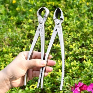 May include: Two silver bonsai pliers with curved blades and long handles. One tool has the text "Grow with love" and the other has the text "Funny". The pliers are held against a backdrop of green foliage and pink flowers.