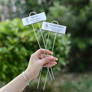 May include: Two metal garden markers, one labelled "BASIL" and the other "ROSEMARY". The basil marker reads "Emma's Garden 04.18.2026" with a floral graphic. The rosemary marker reads "Grandma's Herbs Spring 2026" with a plant graphic.