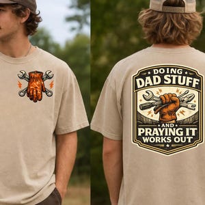 Puede incluir: Camiseta color beige con un diseño gráfico de un guante de trabajo y llaves en la parte delantera. La parte trasera de la camiseta presenta un diseño con el texto "DOING DAD STUFF AND PRAYING IT WORKS OUT" y una mano sosteniendo herramientas.