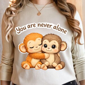 May include: A beige sweatshirt featuring a graphic of two hugging cartoon monkeys. One monkey is orange, the other is brown. The text "You are never alone" is arched above the monkeys in a brown font.