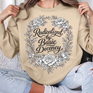 Pode incluir: Camisola bege com a frase "Radicalized by Basic Decency" num design de coroa de flores. O gráfico é em preto e branco, com elementos florais e de folhas detalhados. A camisola é de cor neutra.