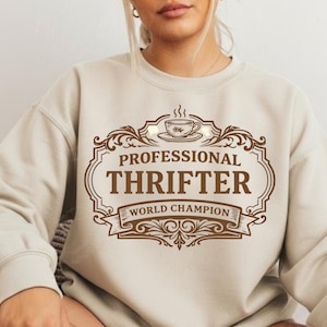 Peut inclure: Sweat-shirt beige avec un motif marron indiquant "PROFESSIONAL THRIFTER WORLD CHAMPION". Le motif comprend une tasse à café et des volutes décoratives. Le sweat-shirt a des manches longues et un col rond.