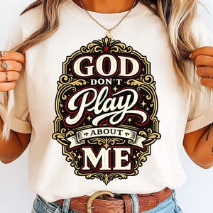 May include: Cream-colored t-shirt featuring a decorative design with the text "GOD DON'T PLAY ABOUT ME". The design is in black, red, and gold, with ornate scrollwork. The shirt has a classic crew neck.