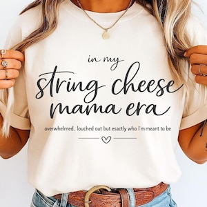 May include: A cream-coloured t-shirt with the phrase "in my string cheese mama era" in black script. The shirt also features the text "overwhelmed, louched out but exactly who I'm meant to be" below the main text, with a heart symbol.