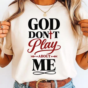 May include: Cream-colored t-shirt with the text "GOD DON'T PLAY ABOUT ME" in black and red. The design includes a cross and arrows. The shirt is paired with blue jeans and a brown belt.