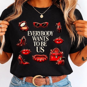May include: Black t-shirt with the text "EVERYBODY WANTS TO BE US" and red illustrations of makeup, accessories, and high heels. The design includes lipstick, sunglasses, and a handbag.