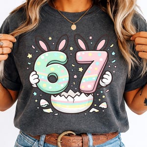 Pode incluir: T-shirt cinzenta escura com um gráfico colorido de Páscoa. O design inclui os números 67, decorados com orelhas de coelho e cores pastel, e uma casca de ovo. A t-shirt é feita de material macio.