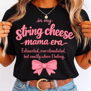 May include: Black t-shirt with the text "in my String cheese mama era" in pink script. Below the text is a pink bow graphic. The shirt also includes the text "Exhausted, overstimulated, but exactly where I belong."