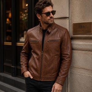 Puede incluir: Chaqueta de cuero marrón con cremallera, dos bolsillos en el pecho y cuello alzado. La chaqueta la lleva una persona con gafas de sol. El fondo incluye un edificio con un letrero que dice "189 COLLINS ST."