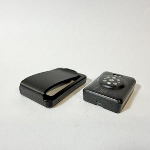 Peut inclure: Deux appareils électroniques noirs sur une surface blanche. L'un est un clip rectangulaire avec un dessus incurvé. L'autre est un petit appareil rectangulaire avec un motif circulaire sur le dessus et un petit port sur le côté.