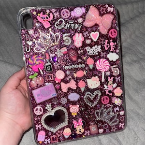 May include: A clear tablet case with a glittery purple background, adorned with various pink and silver charms. The charms include a bow, hearts, Minnie Mouse, a crown, and the word "Love".