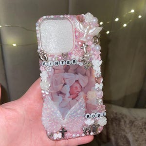 Può includere: Una custodia per telefono rosa decorata con perle, croci, ali d'angelo e i nomi "WINTER" e "ROSE". La custodia presenta una foto di un bambino e vari ciondoli. La custodia è progettata per uno smartphone.