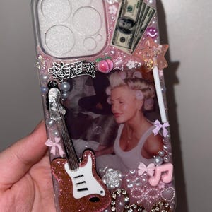 Può includere: Custodia per telefono rosa tempestata di strass con una foto di una donna con i bigodini, una chitarra glitterata, banconote e abbellimenti decorativi. La custodia è adornata con perle, fiocchi e note musicali.