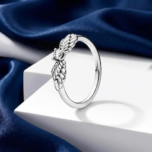 Op de afbeelding: Een zilveren ring met een centrale heldere edelsteen en decoratieve, veerachtige accenten. De ring wordt gepresenteerd op een witte ondergrond met een blauwe satijnen achtergrond, die het ontwerp en de glans benadrukt.