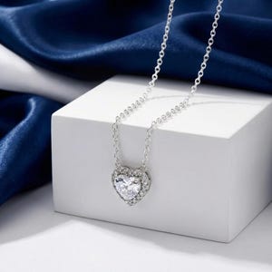May include: A silver necklace featuring a heart-shaped pendant. The pendant is adorned with clear crystals surrounding a central crystal. The chain is delicate, set against a white surface and a blue backdrop.