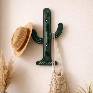 May include: A dark green cactus-shaped wall hook with white accents, holding a straw hat with a brown band and a beige crocheted bag. The decorative hook adds a touch of desert-inspired style to any space.