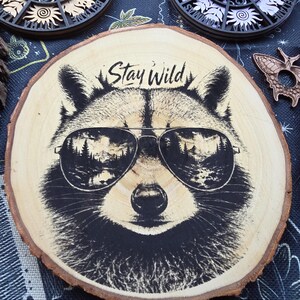 May include: A circular wood slice featuring a black ink illustration of a raccoon wearing aviator sunglasses. The lenses of the sunglasses reflect a mountain landscape with trees. The text Stay Wild is printed in black above the raccoon's head.