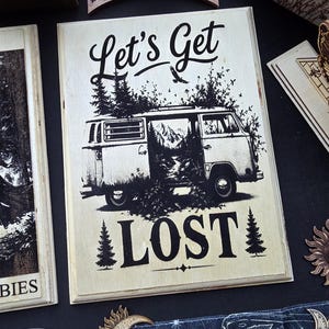 May include: A rectangular wooden plaque featuring a black ink illustration of a vintage camper van surrounded by pine trees and mountains. The text "Let's Get" appears above the van, and the word "LOST" is printed below with pine tree icons.
