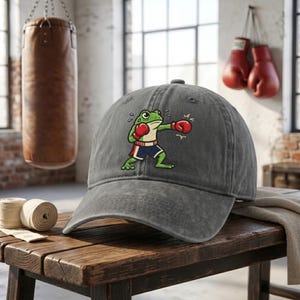 May include: A gray baseball cap featuring a cartoon frog in boxing attire. The frog is green with red boxing gloves and blue shorts. The cap is sitting on a wooden surface, with boxing equipment in the background.