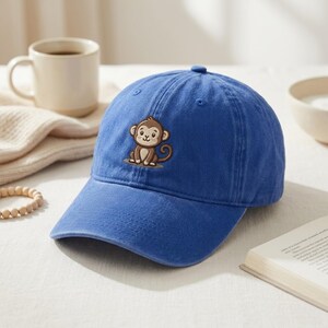 May include: A blue baseball cap with a curved brim and a small, embroidered monkey design on the front. The monkey is brown and tan. The cap is made of a soft, textured fabric.