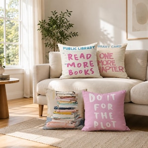May include: Four square throw pillows featuring book-themed designs. Two pillows have cream backgrounds with blue and pink text reading PUBLIC LIBRARY, READ MORE BOOKS, LIBRARY CARD, and ONE MORE CHAPTER. One shows a stack of books, one is pink with white text DO IT FOR THE PLOT.