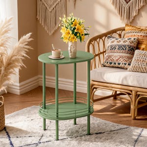 May include: An oval-shaped sage green metal side table with two tiers. The top tier holds a white ceramic face-shaped vase filled with yellow lilies and a small white mug on a saucer. The bottom tier features a thin metal railing around the edge.