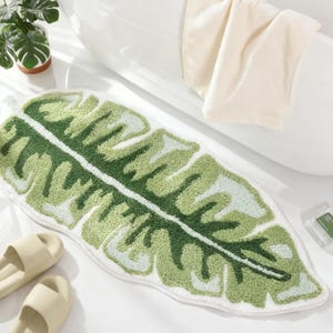 May include: A green and white leaf-shaped bath mat. The mat features a detailed design with varying shades of green and white, resembling a tropical leaf. The mat is placed near a white bathtub and a pair of beige slides.