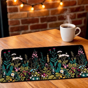 May include: A rectangular desk mat featuring a black background with a vibrant botanical print of wildflowers in shades of pink, white, yellow, and teal green. The mat has rounded corners and is positioned on a wooden surface next to a white mug.