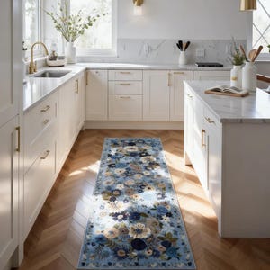 May include: A rectangular runner rug featuring a light blue background with a dense, multi-colored floral pattern in shades of navy blue, cream, beige, and sage green. The rug is positioned on a wooden floor between white kitchen cabinets.