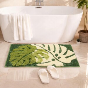 May include: A green and cream-colored bath mat with a large leaf design sits in front of a white bathtub. Two white slippers are placed on the floor. A white towel hangs on the side of the tub. A potted plant is in the background.