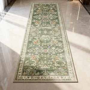 May include: A rectangular, green runner rug with an intricate floral pattern. The rug features a central design of flowers and leaves in shades of orange, blue, and white, framed by a cream-colored border. The rug is placed on a light-colored floor.