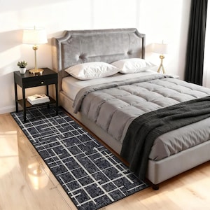 May include: A rectangular, dark blue rug with a white grid pattern is placed in front of a bed. The rug is approximately 45 inches long. The bedroom features a gray bed frame, headboard, and bedding, with a black nightstand and gold lamp.